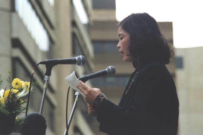 You view a young woman of Asian descent speaking into a microphone on a stage decorated with yellow flowers on an overcast day.