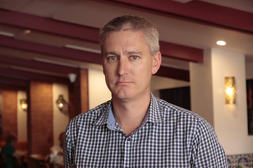 A man in a collared shirt, standing inside a venue, looking serious.