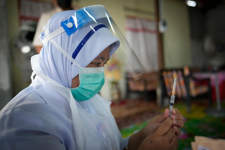 A nurse in a surgical mask and a face mask holds a syringe.