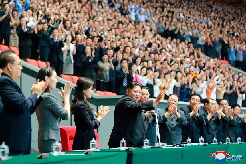 The entire crowd of a large stadium look at Kim Jong-un who waves while standing.