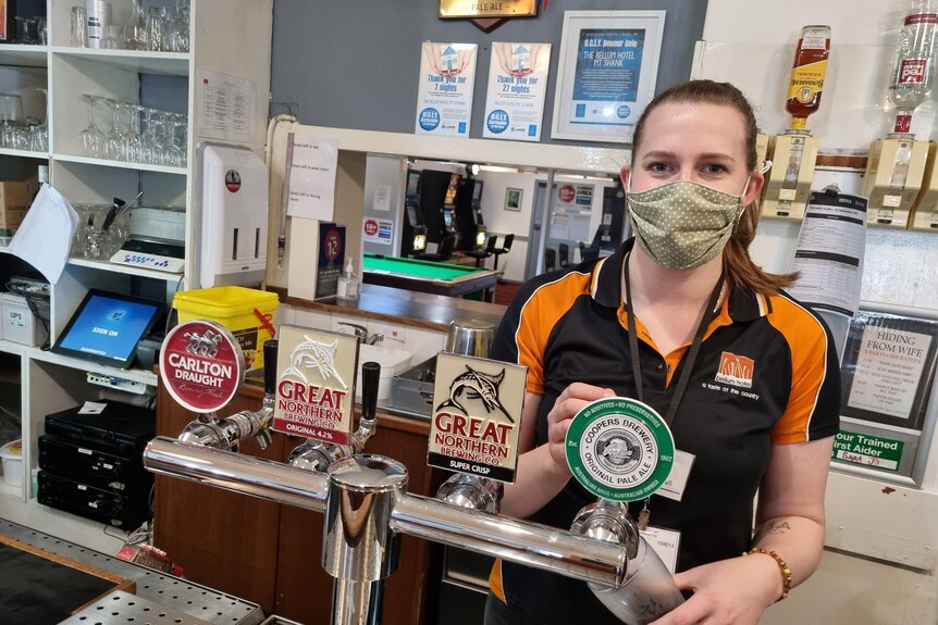 A woman stands behind a bar counter, wearing a mask and pouring a beer