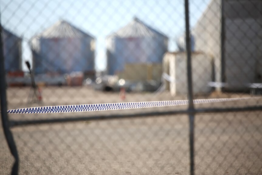 Police tape around a fence, with large grain silos in the background. 