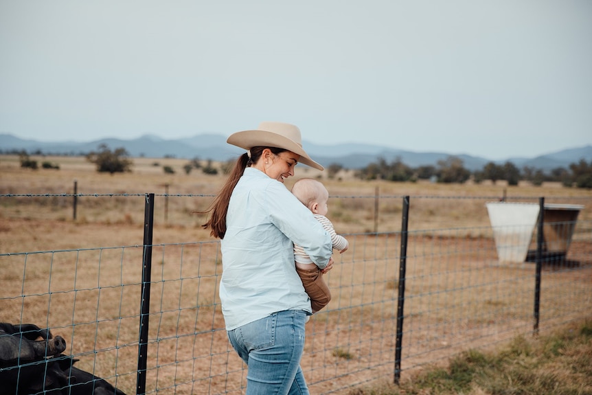 A woman stands in a paddock holding a baby.