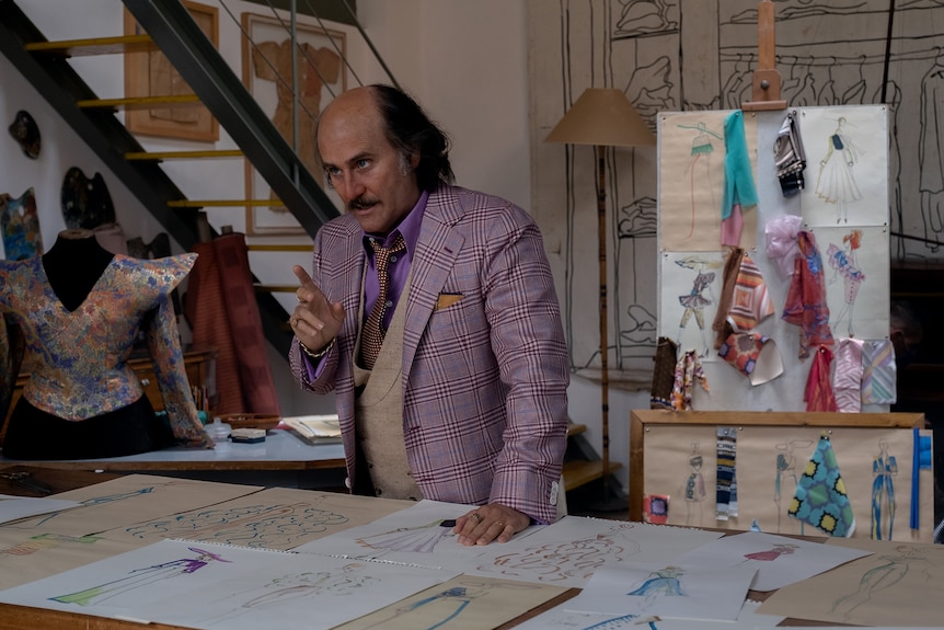 Bald Italian man in checkered purple three-piece suit leans over the table covered in clothes design sketches