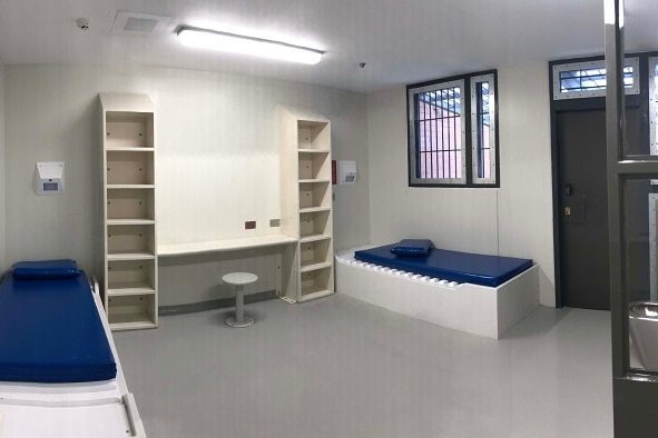An empty prison cell lit by fluorescent light, contains two single beds, shelves, a desk and stool.
