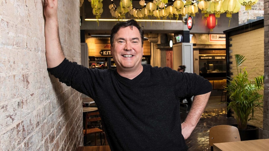 A man with short dark hair, wearing a dark jumper, leaning against a brick wall in a cafe.