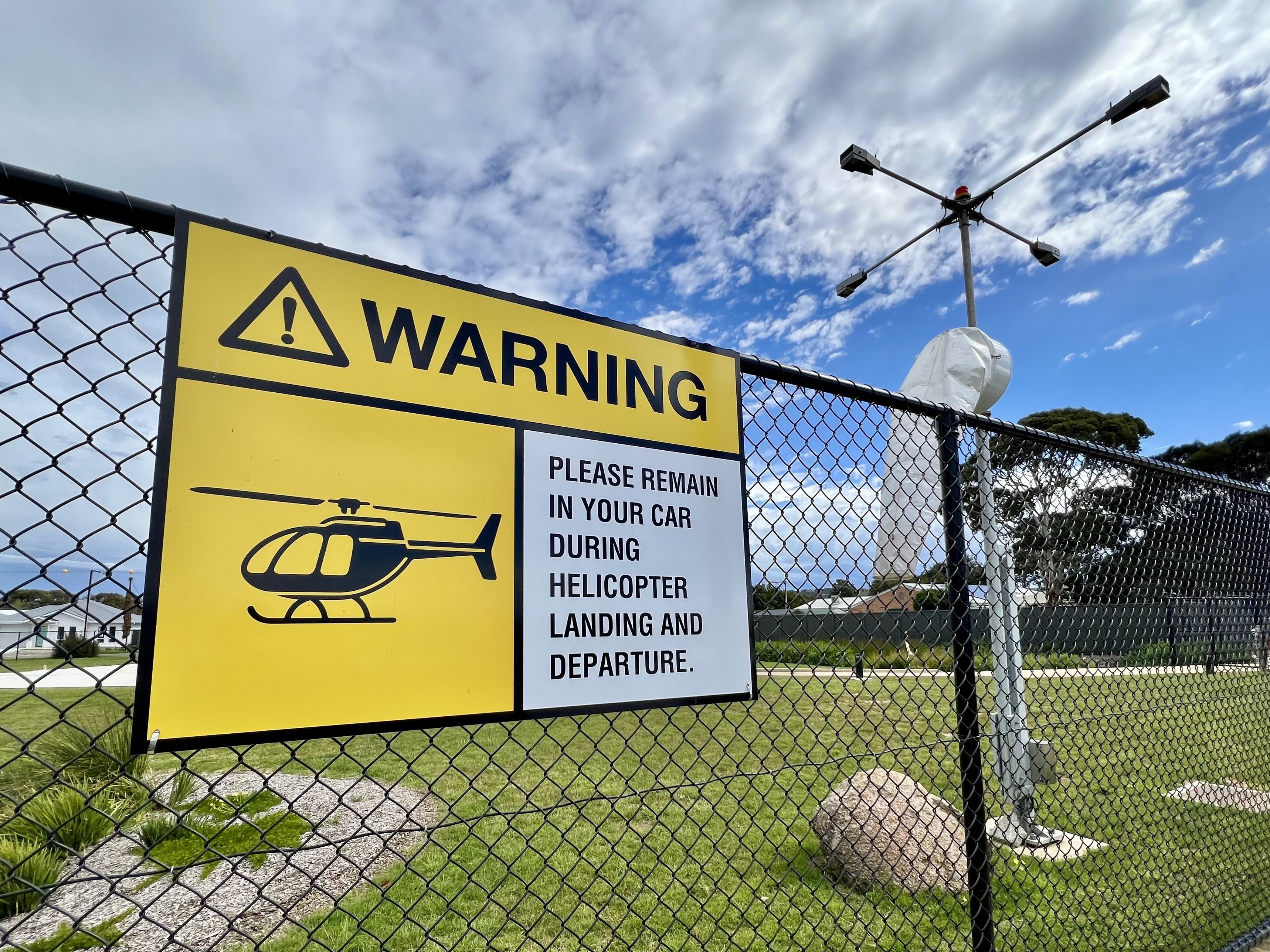 A fence with a sign warning of helicopter arrivals with a windsock behind.
