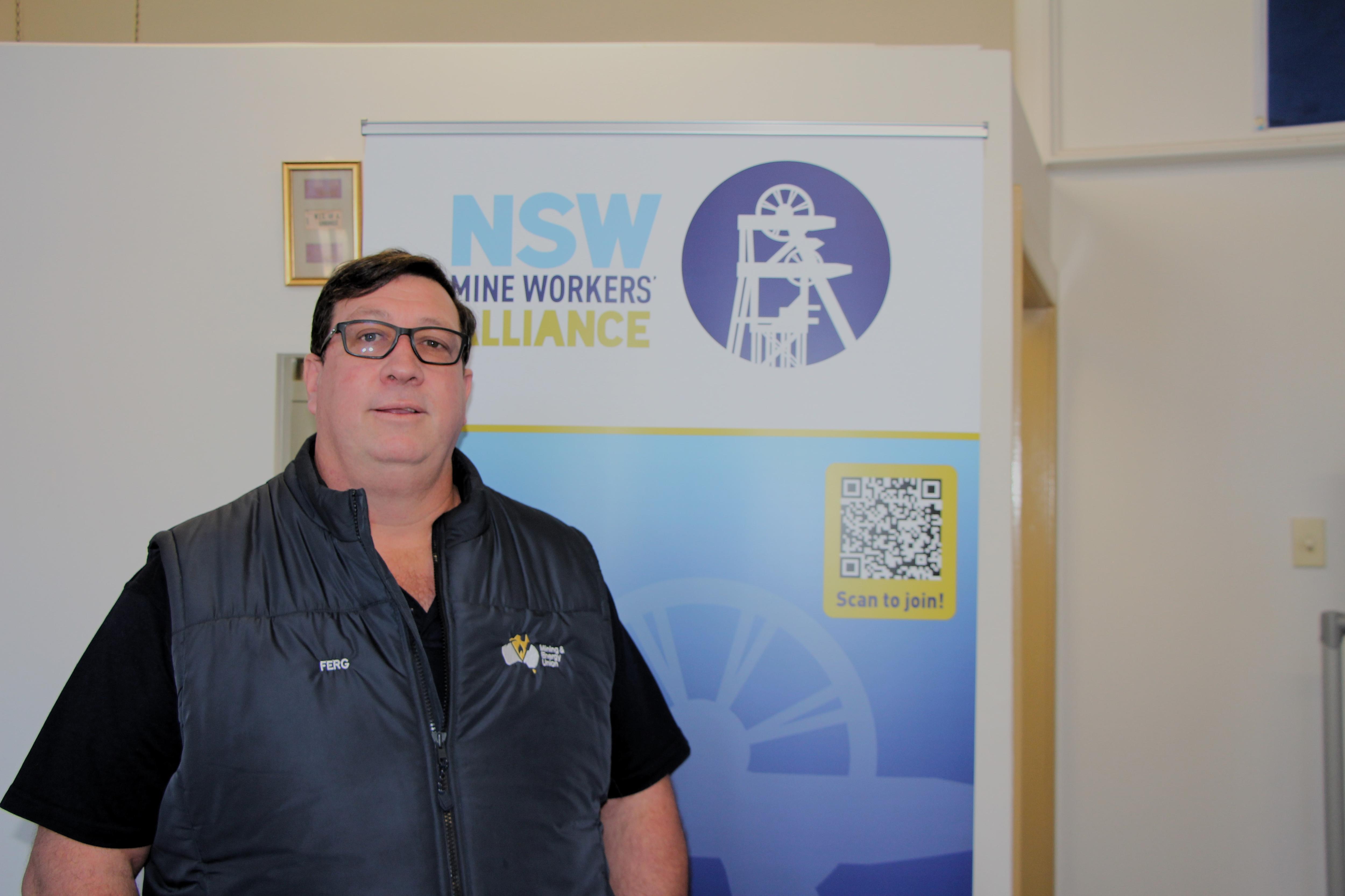 Un hombre está parado frente a un cartel de la Alianza de Trabajadores Mineros de Nueva Gales del Sur.