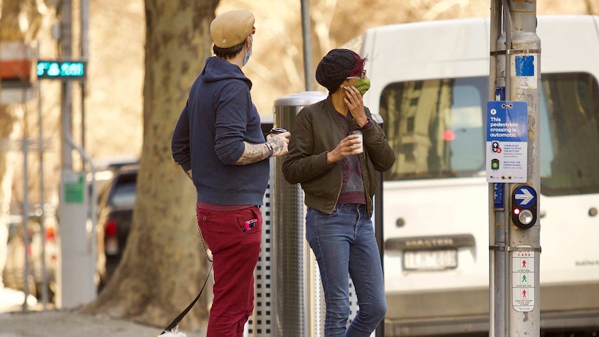 Two people wearing masks, with a small dog in a yellow jacket, standing at a quiet pedestrian crossing in Melbourne.