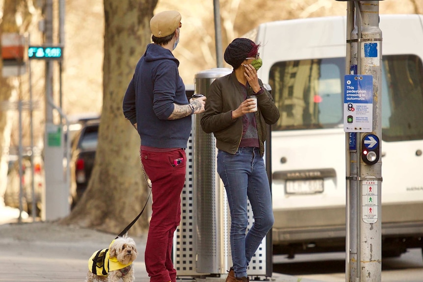 Two people wearing masks, with a small dog in a yellow jacket, standing at a quiet pedestrian crossing in Melbourne.