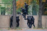 Nine armed police in helmets and protective gear climb a brick fence