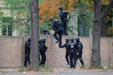 Nine armed police in helmets and protective gear climb a brick fence