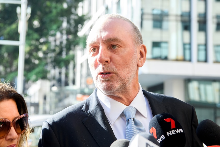 a man wearing a suit and tie talking at a reporter on a bright sunny street