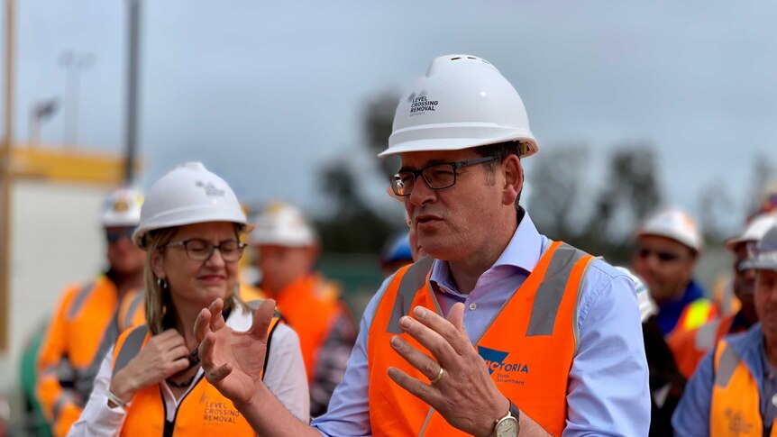 A man in a blue shirt and orange high-vis jacket and hard hat holds his hands out as he addresses the media.