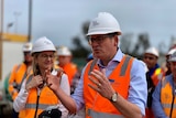 A man in a blue shirt and orange high-vis jacket and hard hat holds his hands out as he addresses the media.