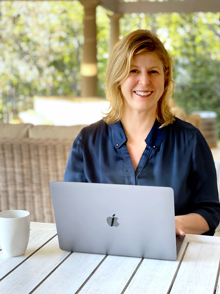 A middle-aged woman with blonde hair and a navy shirt sits at a laptop and smiles at the camera