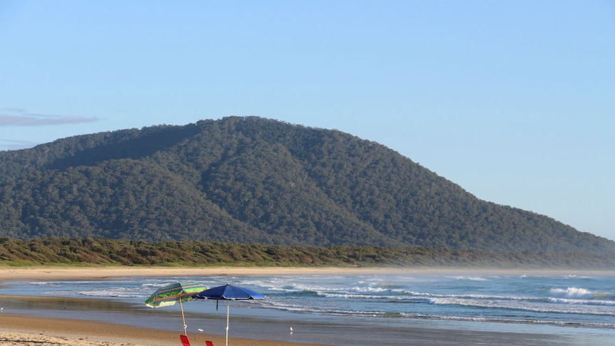 Two red deck chairs and umbrellas on a beach, with a mountain and blue sky in the background.