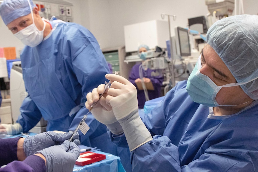 A doctor in blue hospital scrubs and face mask reaches for surgical equipment watched by colleagues.