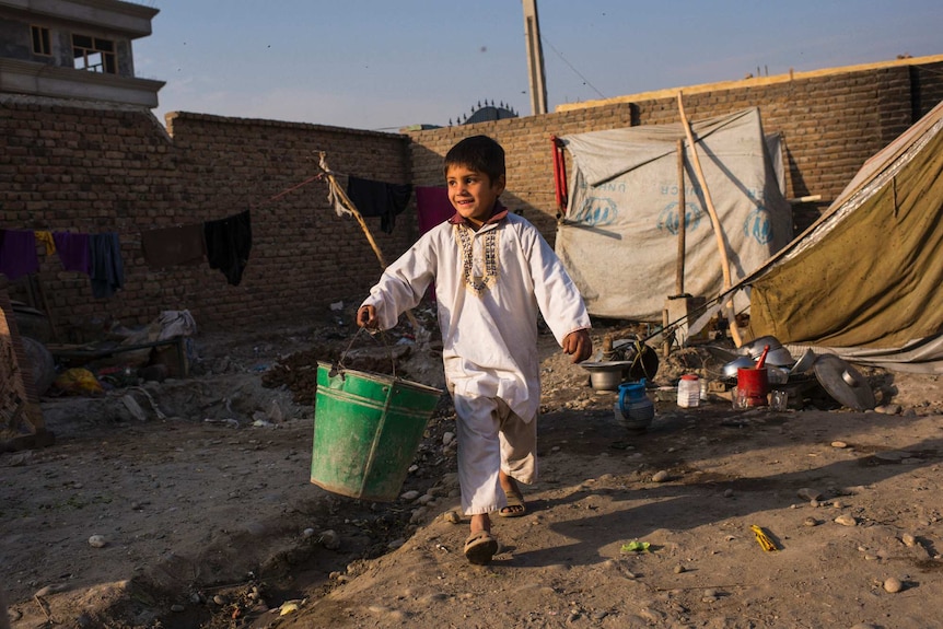 A young Afghan boy carries a bucket through a run-down camp in eastern Afghanistan.