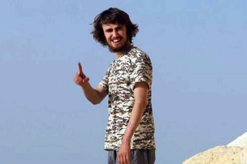 A man with shaggy hair and a camo t-shirt holds up his index finger with desert in the background.