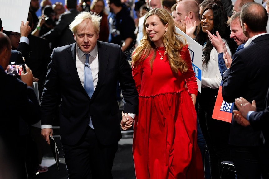 Boris Johnson holds hands with his wife Carrie, who is wearing a billowing red dress.