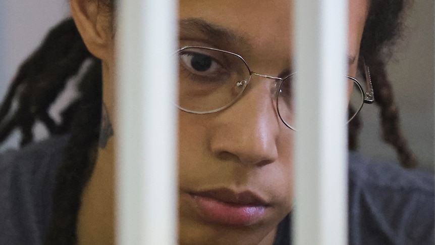 A close-up of a solemn Black woman wearing wire-framed glasses through prison bars