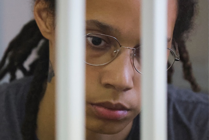 A close-up of a solemn black woman wearing wire-framed glasses through the prison bars