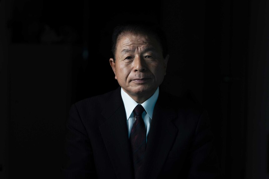 A Japanese man in a suit with only his face visible in the darkness