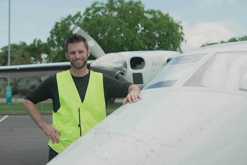 A man leans against a small plane, he is wearing a high-vis yellow vest 