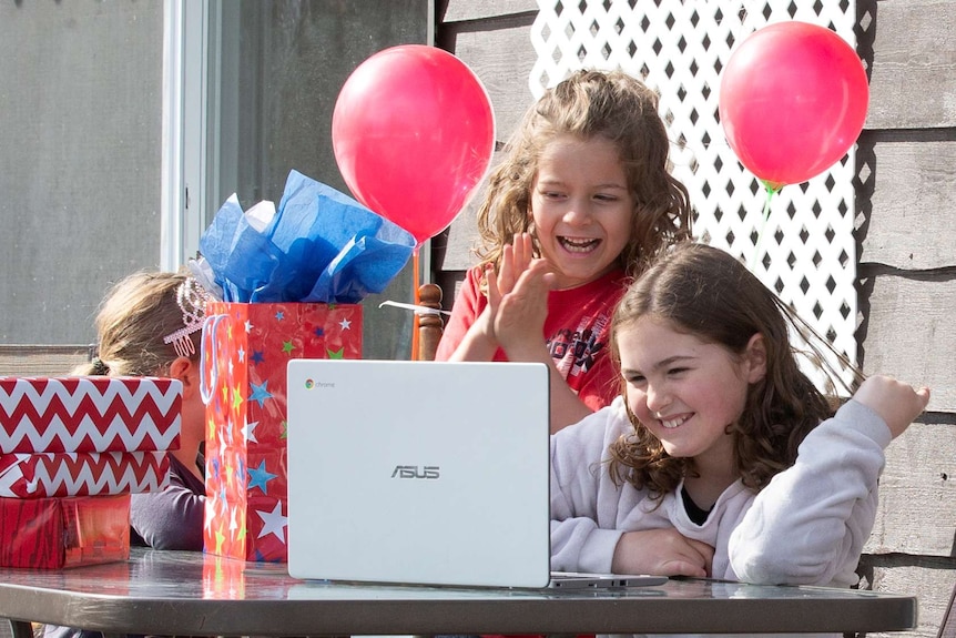 Two children laugh and smile at a computer screen during a birthday party surrounded by balloons and gifts