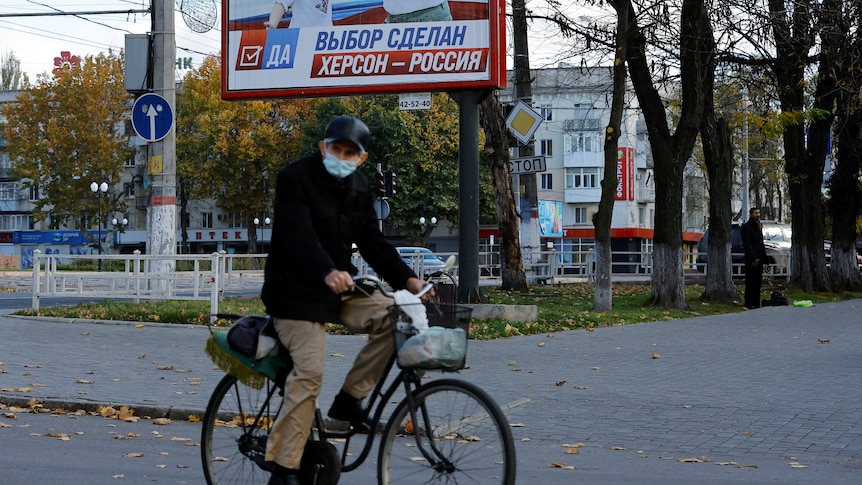A street scene, with an older man riding a bike, with a blue, red and white billboard behind him.