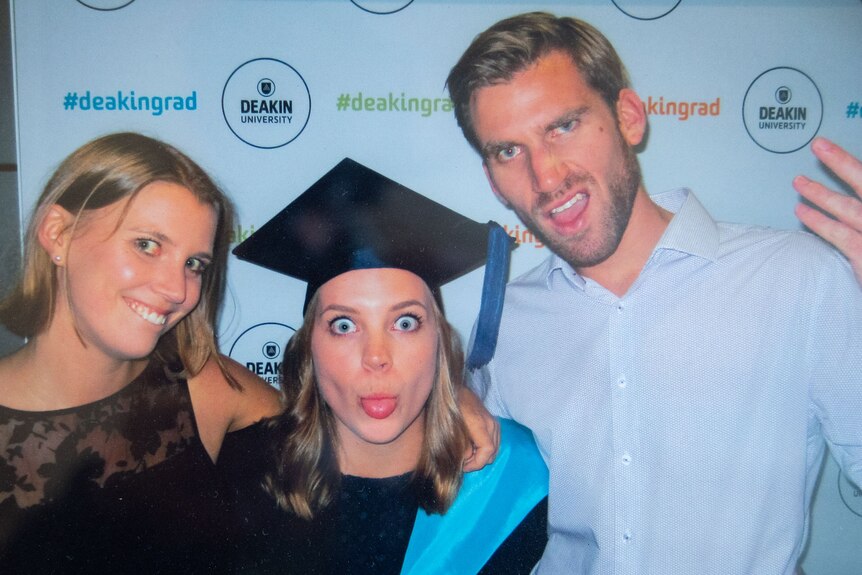 Young woman in mortarboard hat sticks tongue out, clebrating graduation with brother and sister