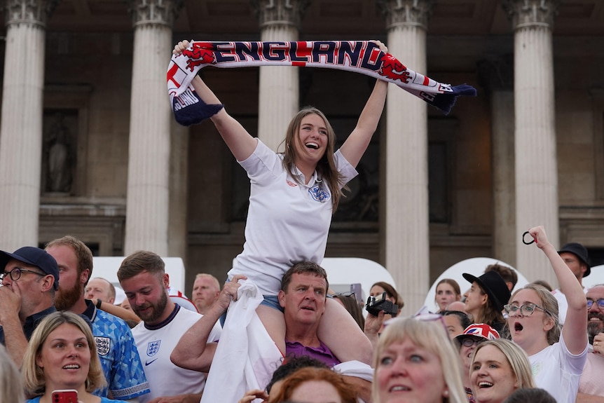 A woman is sitting on a man's shoulders in a crowd. She holds a scarf above her head with "England" written on it. 