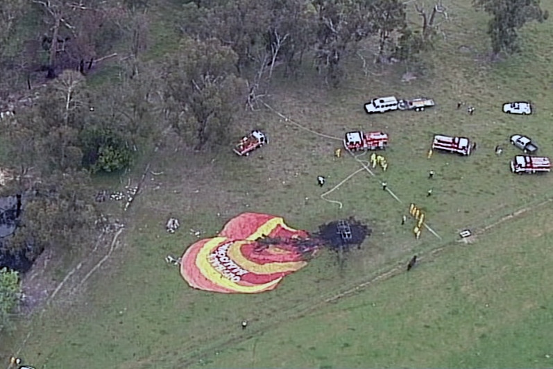 An aerial shot showing the crash of the balloon, with charred area where the basket is, in a paddock. There are vehicles nearby.