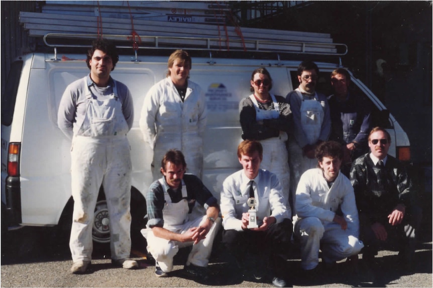 A grainy photo of nine men wearing white overalls and jumpsuits, posing for a photo in front of a van.