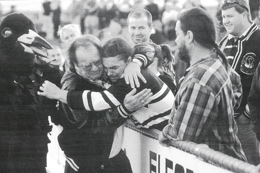 A rugby league coach hugs a fan as the team mascot looks on. 