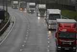 Trucks drive down a highway on a grey overcast day with their headlights on.