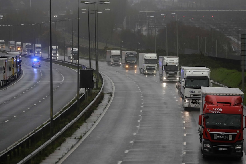 Trucks drive down a highway on a grey overcast day with their headlights on.