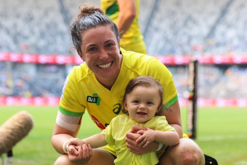 Emilee Cherry crouches down and holds helps her daughter stand on the rugby field.