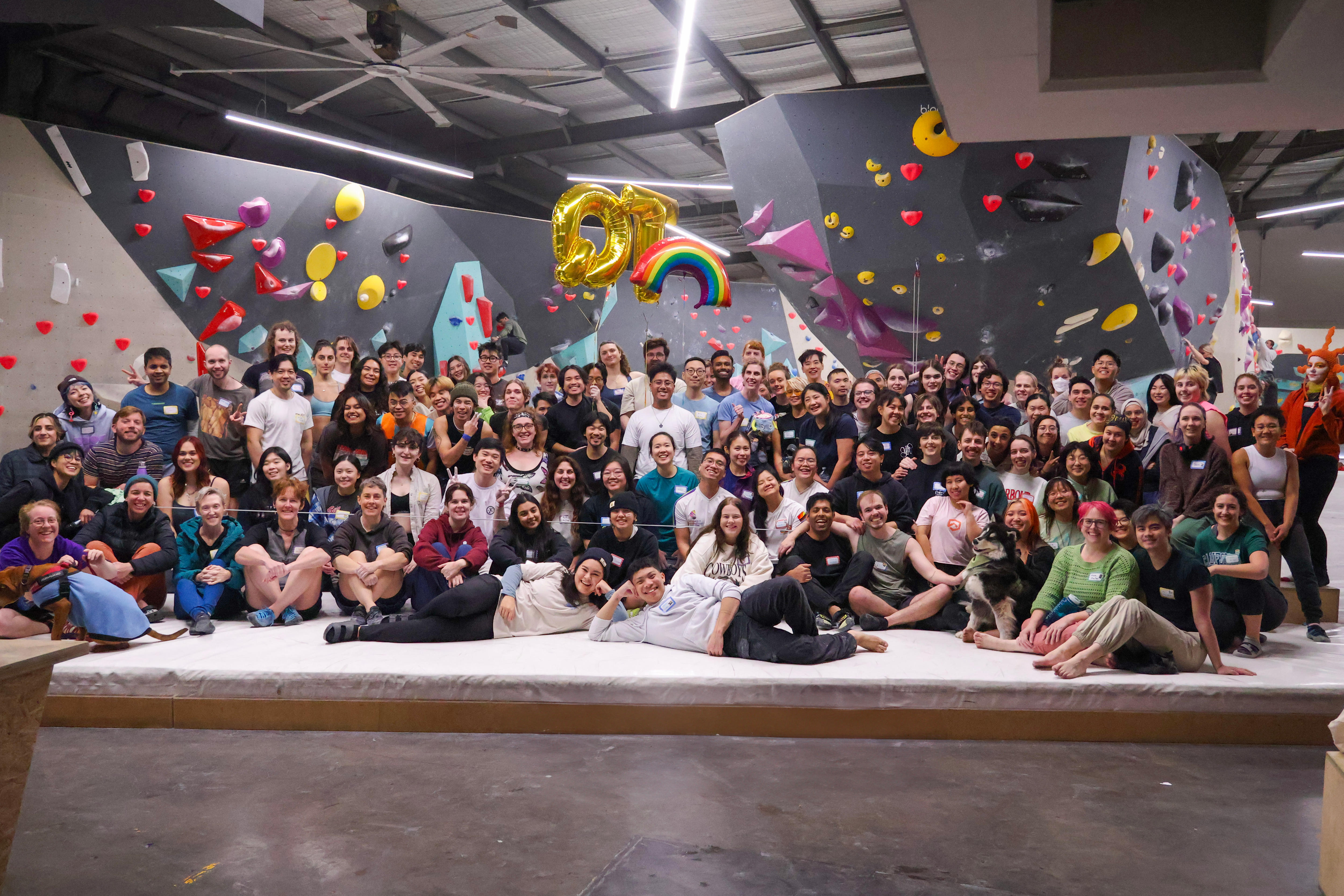A large group shot of social climbers in an indoor rock climbing gym, they're posing, smiling, with a rainbow balloon.