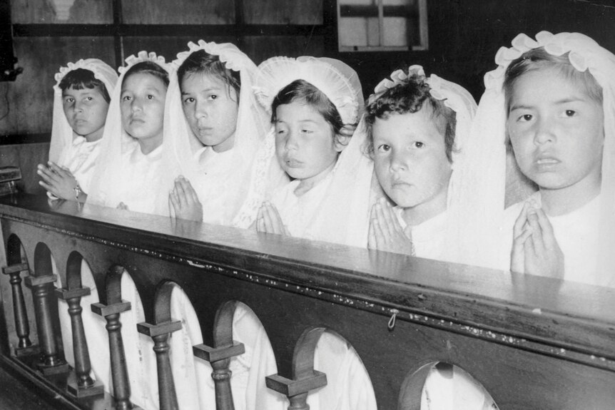 A black and white image of little girls in white communion dresses praying at a pew