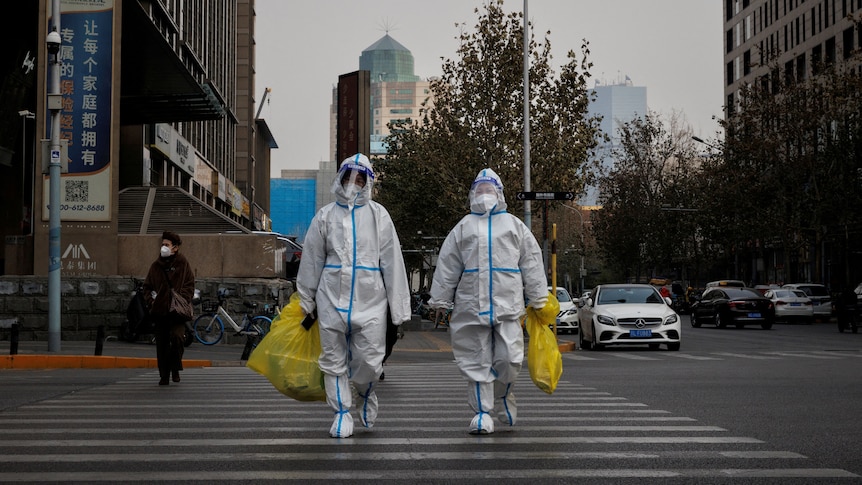 Two people wearing full body protective suits cross a road carrying yellow garbage bags