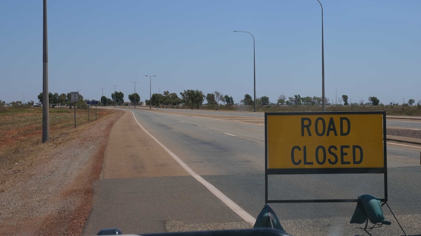 A road closed sign sits on an empty road beneath a bright blue sky.