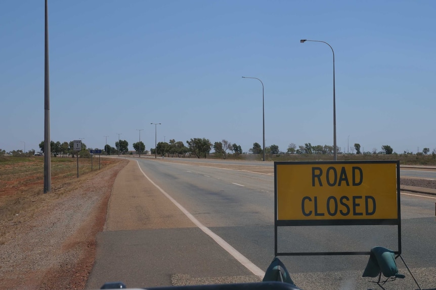 A road closed sign sits on an empty road beneath a bright blue sky.