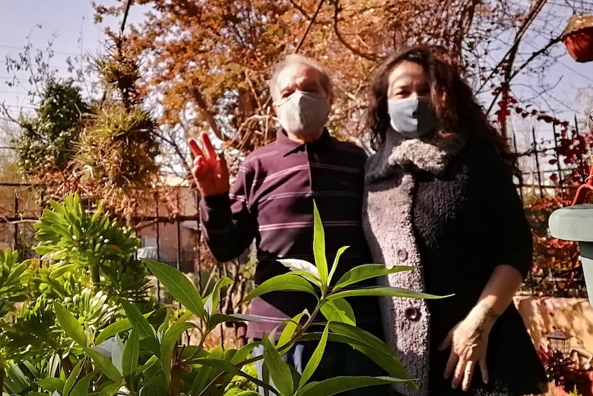 A man and a women stand in a sunny garden wearing face masks.