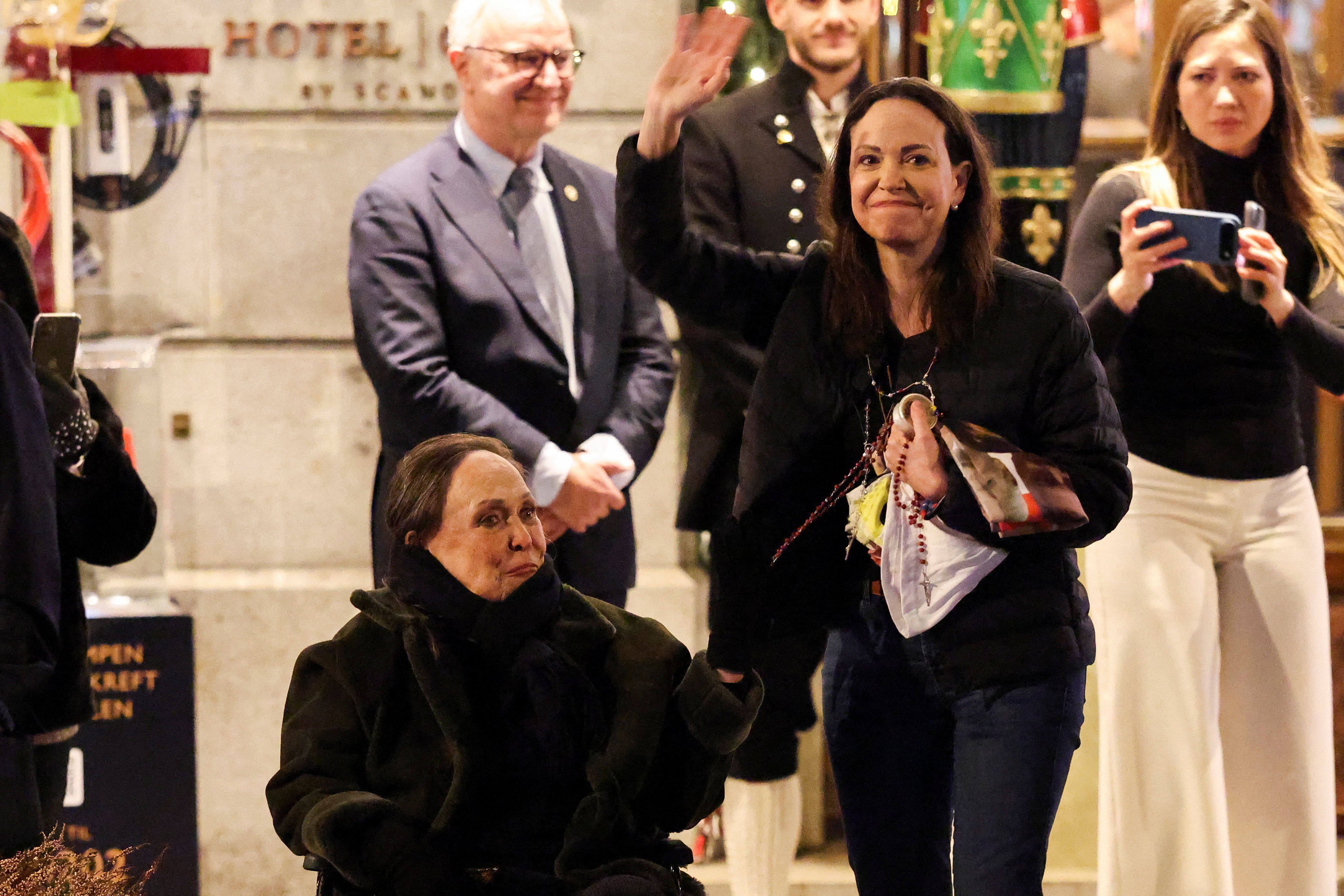 María Corina Machado, vestida con jeans oscuros y una chaqueta acolchada negra, sonríe y saluda junto a su madre, quien está sentada.