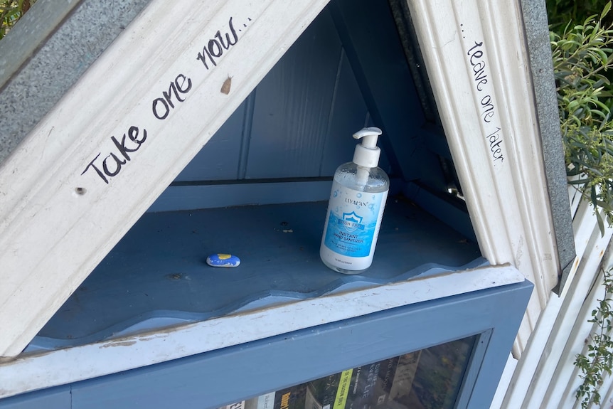 Hand sanitizer can be found at the top of a free bookcase, with an inscription on the cabinet saying "take one now, leave one later"
