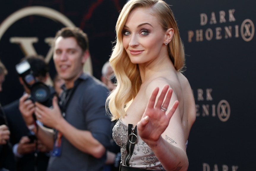 A close up of Sophie Turner smiling while lookng over her shoulder while a man with a camera looks on.