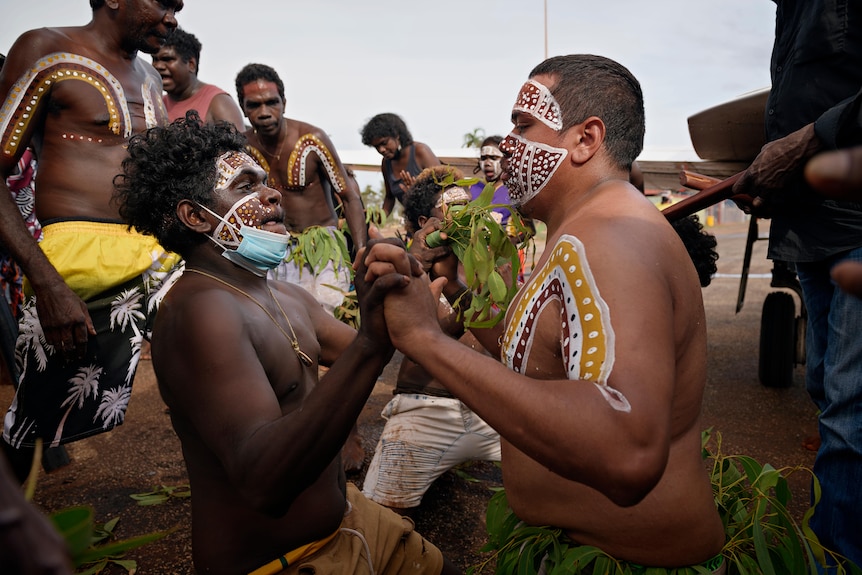 Two men with painted faces and bodies hold hands. They look serious.