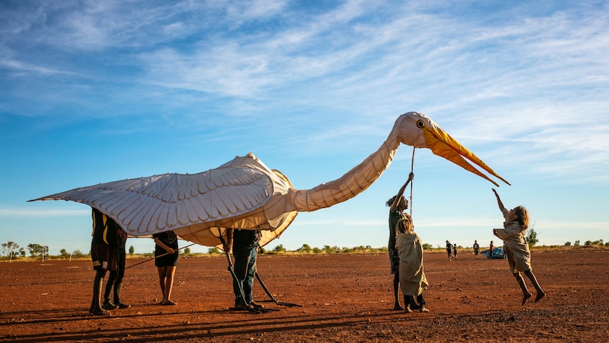 A small child jumps to touch the long beak of a giant puppet bird, surrounded by red dirt and blue sky